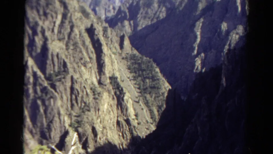 1967 Colorado 8mm frame showing rugged canyon cliffs, evergreen patches, and strong sunlight against dark shadowed rock.