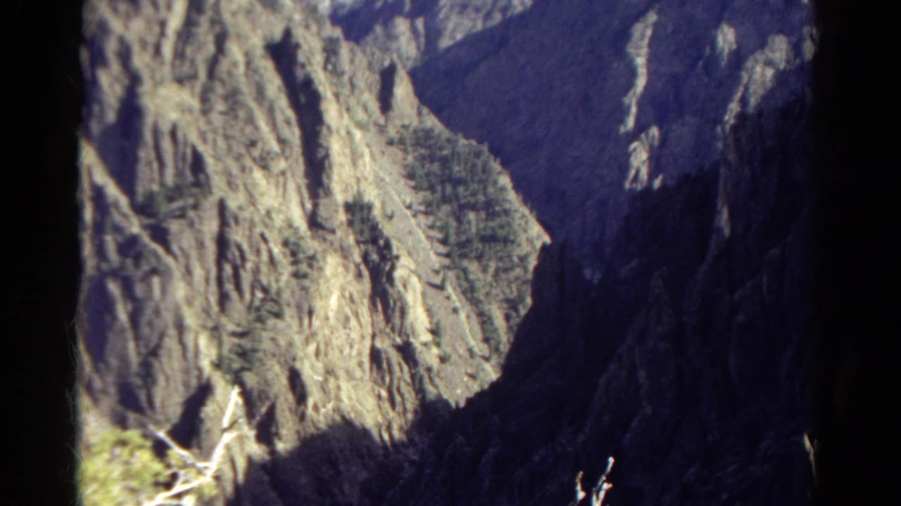 Colorado mountain vista on 8mm film, with steep rock faces, scattered evergreens, and a dark canyon wall at right.