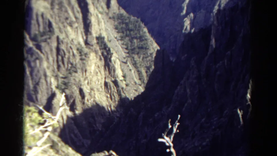 Final 1967 Colorado home-movie frame showing a shadowed canyon wall, bright cliff face, sparse trees, and pale branches in the foreground.