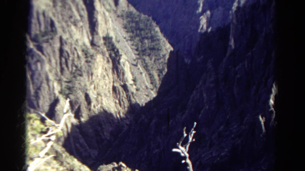 1967 Colorado 8mm frame showing a steep sunlit rock face, deep shadowed canyon, and sparse foreground vegetation from a mountain viewpoint.