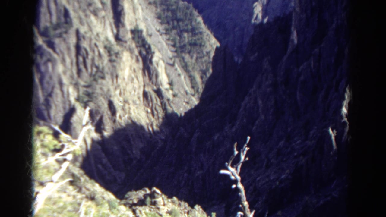 1967 Colorado mountain view on 8mm film, with pale cliff textures, deep blue-black shadow, rocky terrain, and a bare branch in the foreground.