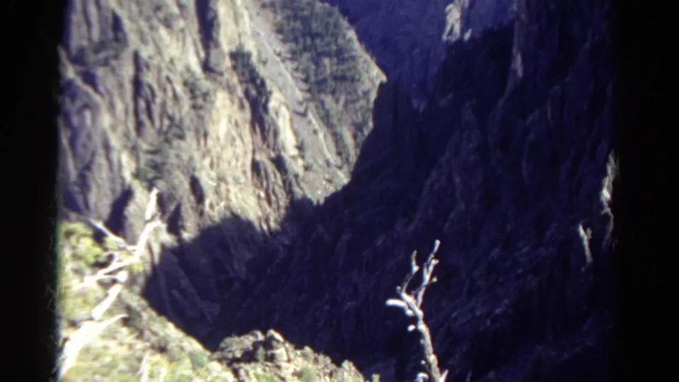 1967 Colorado mountain view on 8mm film, with pale cliff textures, deep blue-black shadow, rocky terrain, and a bare branch in the foreground.