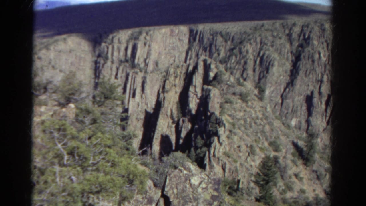 1967 Colorado 8mm frame showing steep canyon rock faces, shadowed vertical formations, evergreen trees, and scrub vegetation.