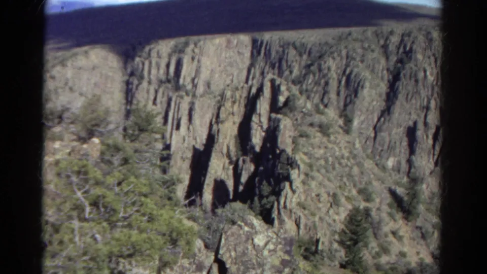 1967 Colorado 8mm frame showing steep canyon rock faces, shadowed vertical formations, evergreen trees, and scrub vegetation.