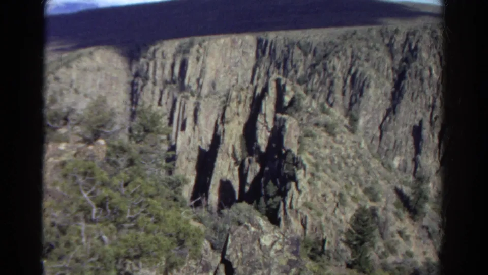 1967 Colorado home-movie frame showing rocky canyon walls, dark vertical formations, evergreen trees, and dry mountain brush.