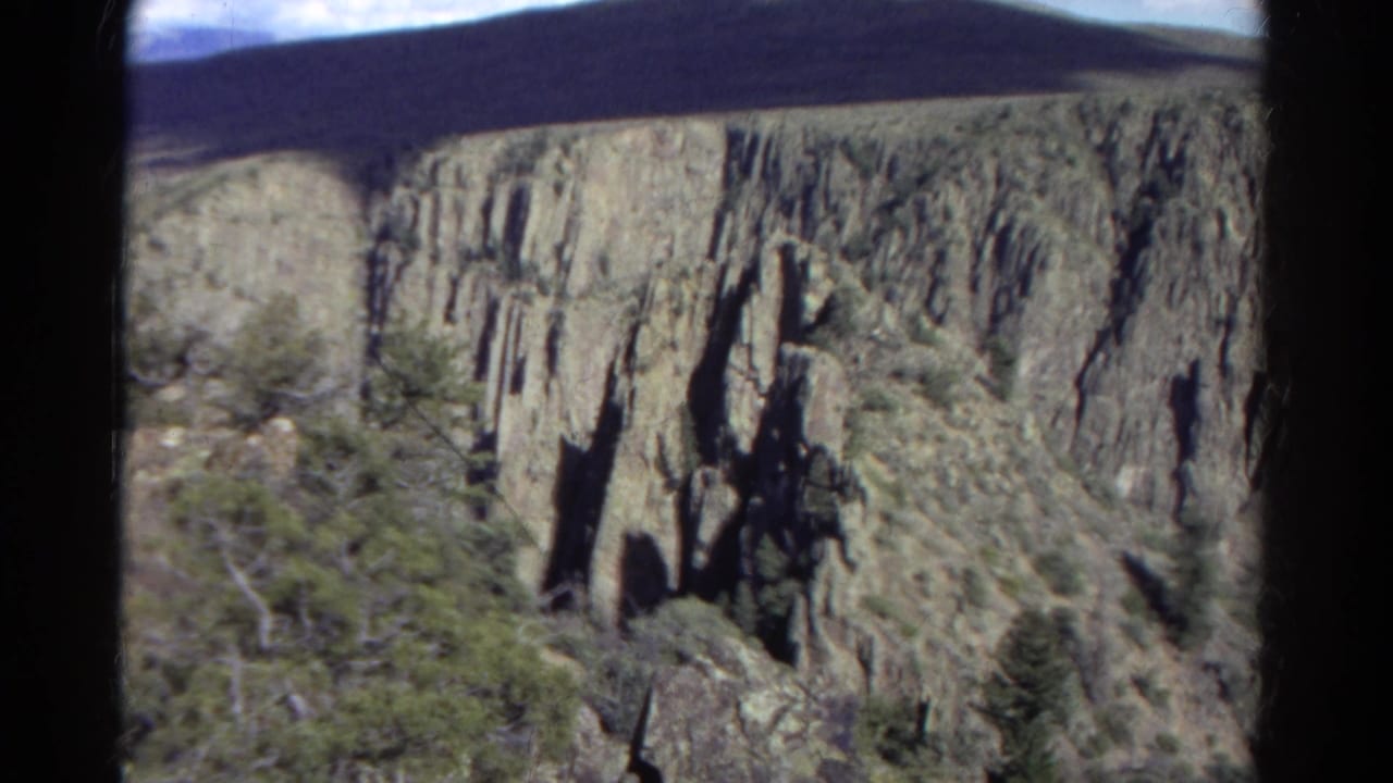 1967 Colorado 8mm frame showing a craggy canyon wall, shadowed rock, sparse shrubs, and rounded black film-frame edges.