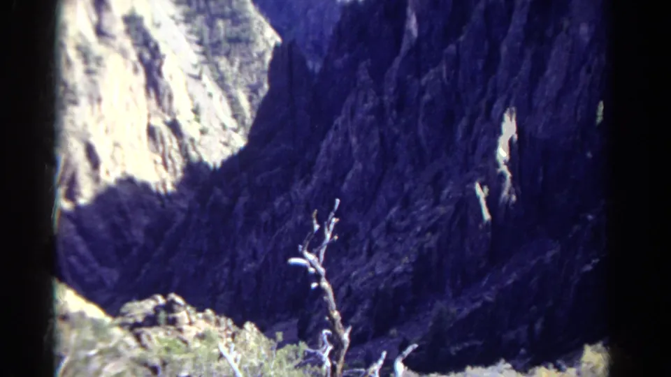 1967 Colorado 8mm frame of a steep canyon wall, with heavy blue shadow, sunlit stone, rocky foreground, and sparse brush.