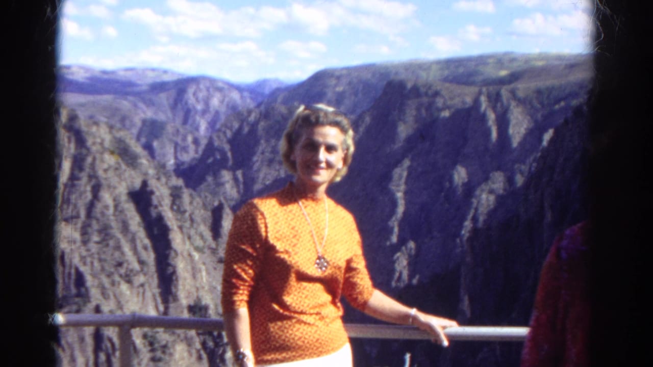 In Colorado, 1967, a woman in an orange patterned top and white pants smiles at a canyon overlook railing.