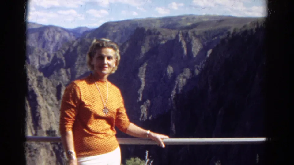 The woman remains near the overlook rail, with shadowed canyon walls and distant Colorado ridges behind her.