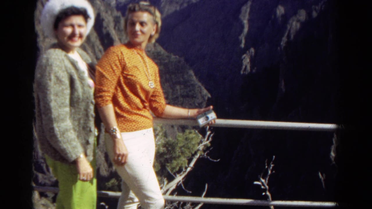 Two women face the camera near a railing at a rocky mountain viewpoint.