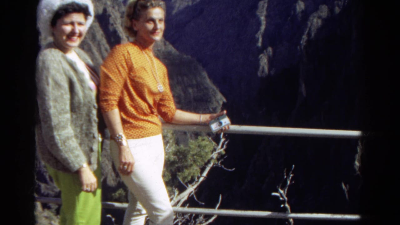 Two adult women in bright 1960s clothing pose at a Colorado mountain overlook beside a metal railing.