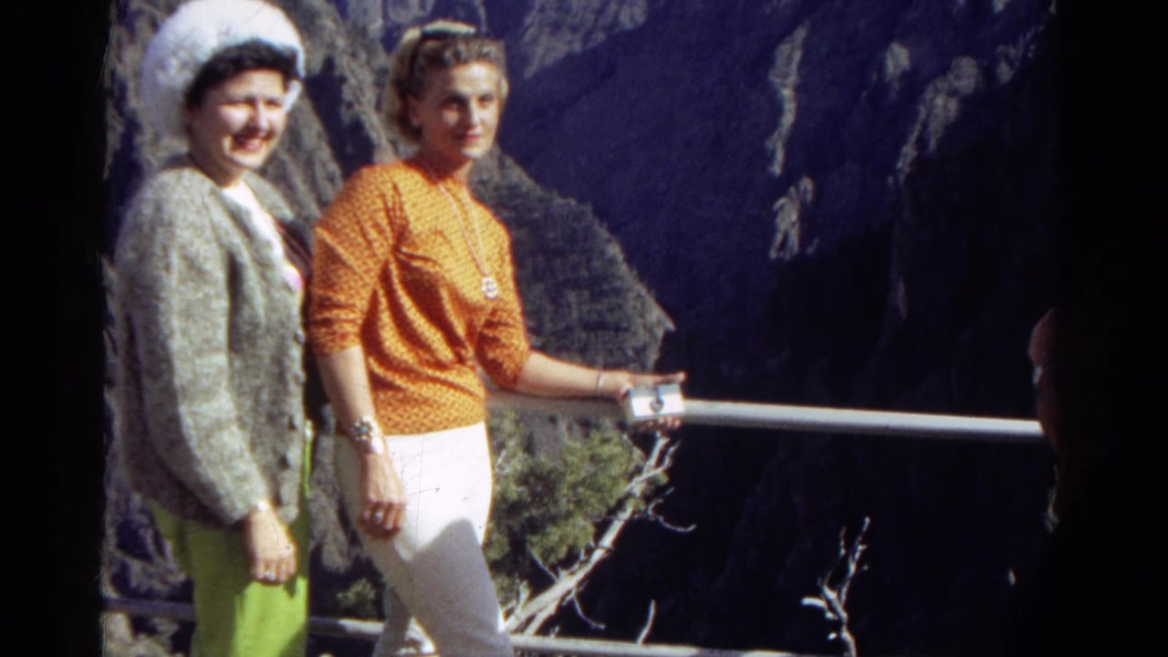 In 1967 Colorado 8mm footage, two dressed-up women stand by a metal overlook railing with dark mountain canyon walls behind them.