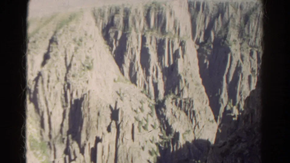 1967 Colorado 8mm frame showing sunlit canyon terrain, steep rock columns, shadowed recesses, and scattered green growth on the slopes.