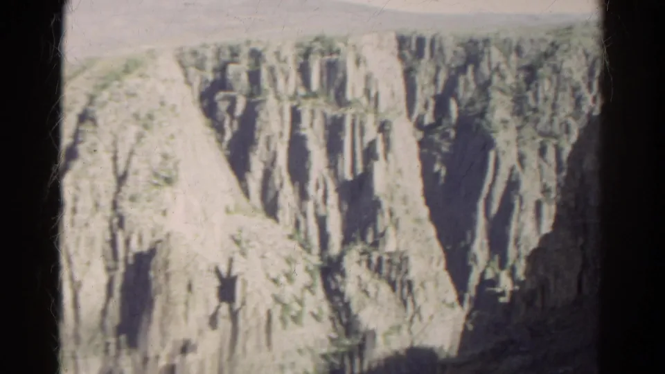 1967 Colorado 8mm frame with a wider canyon view, pale angled slope, vertical cliff formations, dark shade, and sparse greenery.