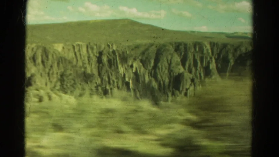 1967 Colorado 8mm travel view from a moving vehicle window, showing steep cliffs, roadside blur, distant hills, and pale sky.