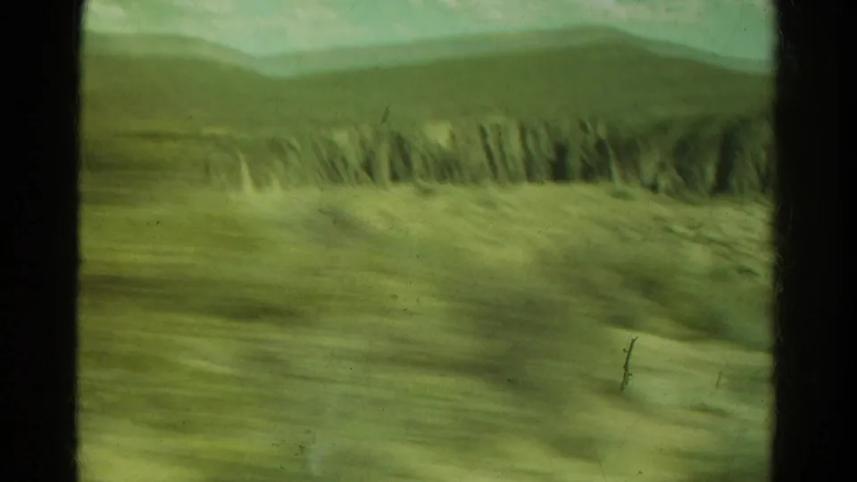 1967 Colorado travel footage frame with motion-blurred ground, distant canyon wall, rounded hills, and dark window framing.