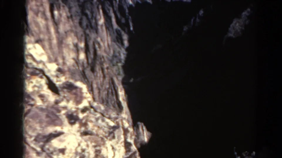 1967 Colorado home-movie frame showing a bright rocky ledge and cliff texture against the deep shadow of a mountain ravine.