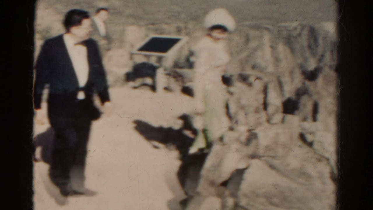 Adults in 1960s dress clothing appear to walk through rocky Colorado mountain terrain in a home-movie frame.