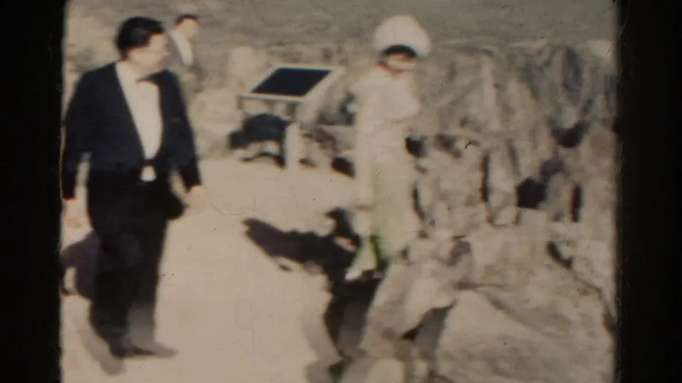 Adults in 1960s dress clothing appear to walk through rocky Colorado mountain terrain in a home-movie frame.