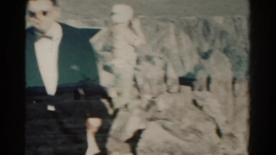 Soft 8mm frame of adults on rocky Colorado terrain, with formal clothing and a still camera visible.