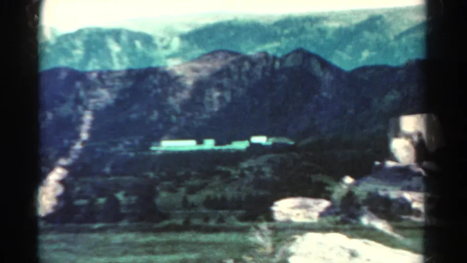 1967 Colorado landscape frame showing a dark ridge, rocky foreground, trees, and a pale feature along the valley floor.
