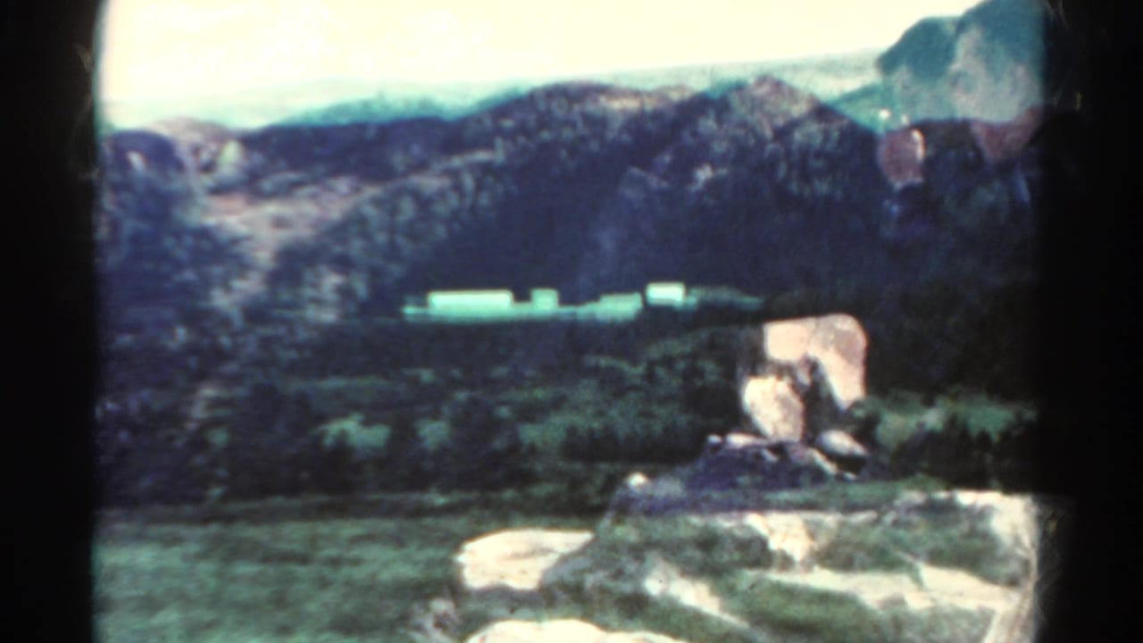1967 8mm home-movie view of rugged Colorado terrain with distant mountains, scrub, and pale roadside forms.