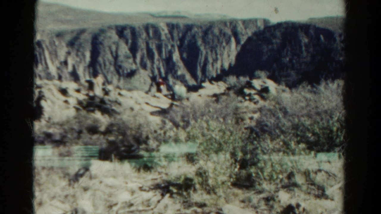 1967 Colorado 8mm frame showing pale desert ground, sparse scrub, and steep rocky cliffs under faded color exposure.
