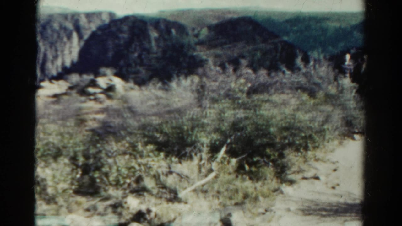 1967 Colorado 8mm view of sparse brush, pale soil, and rugged cliff faces in a visitor-shot desert landscape.