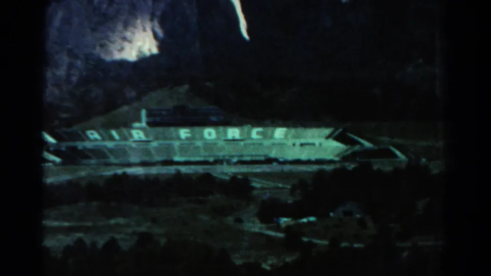 Dark 1967 8mm frame showing a long Air Force-marked structure or sign with high-contrast bright areas above it.