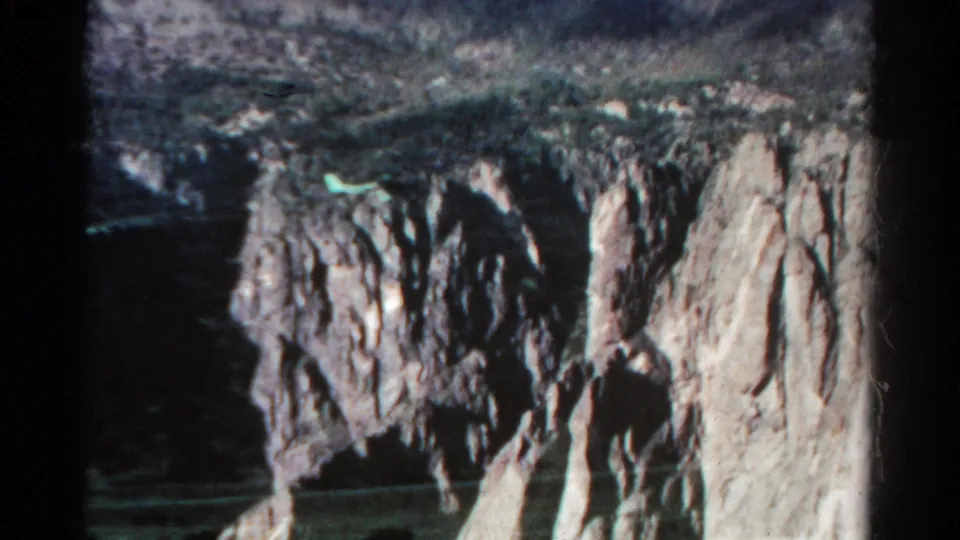 1967 Colorado 8mm frame widening to show rugged rock formations, a deep shadowed opening, and a small greenish reflection against the dark terrain.
