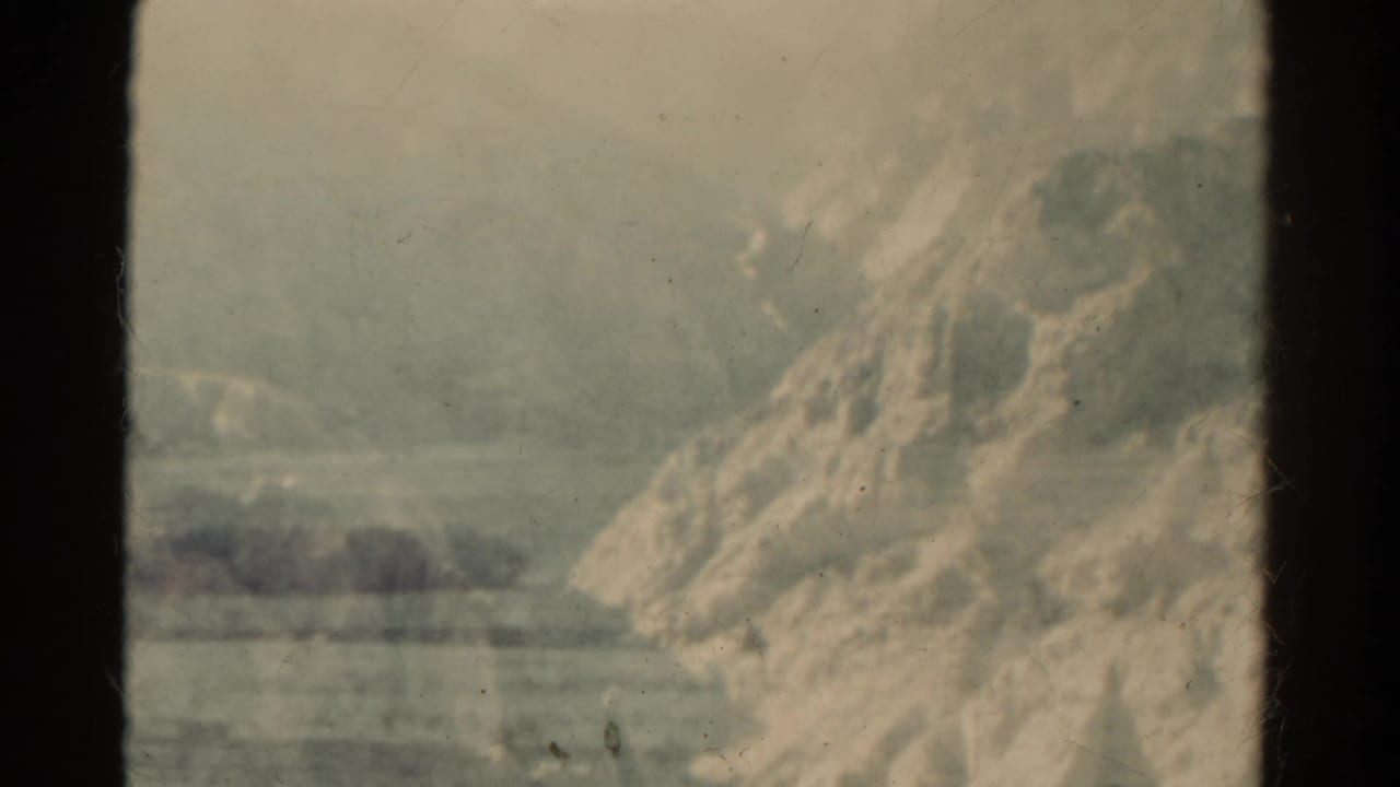 1967 Colorado 8mm frame showing a rocky slope beside a pale open landscape, viewed through dark window edges from a moving vehicle.