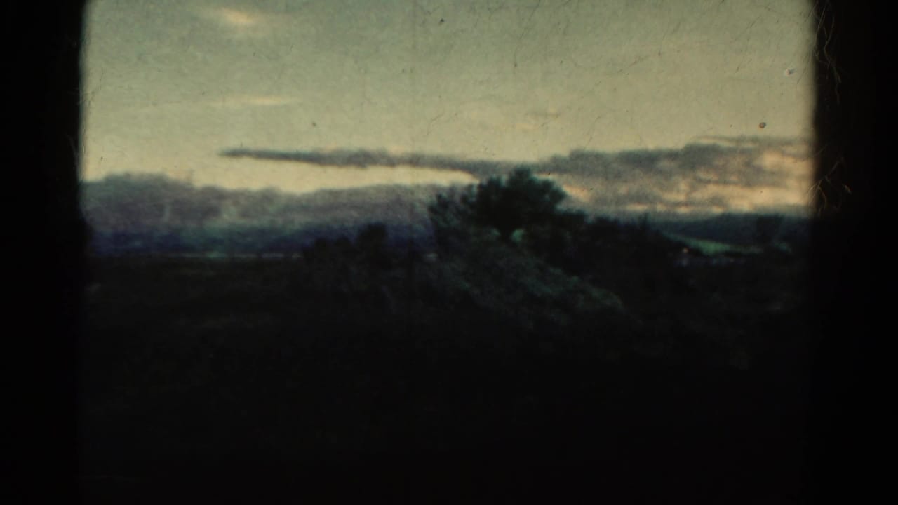 1967 Colorado home-movie frame showing a dim countryside view with distant hills, silhouetted trees, layered clouds, and visible film borders.