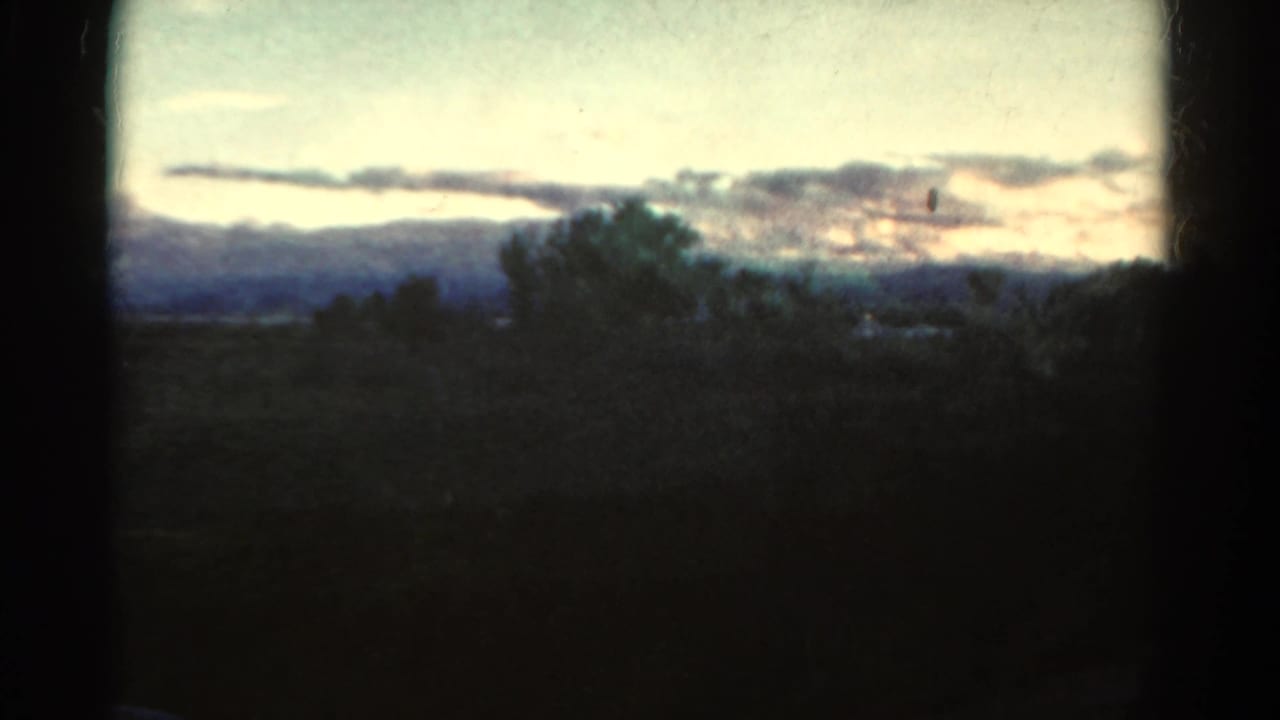 1967 Colorado countryside view with shadowed trees, low hills, horizontal clouds, pale evening light, and dark film gate borders.
