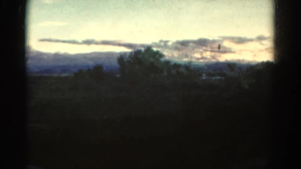 1967 Colorado countryside view with shadowed trees, low hills, horizontal clouds, pale evening light, and dark film gate borders.