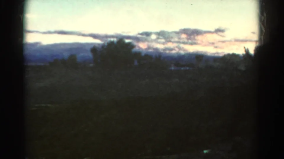 1967 Colorado 8mm frame showing a dark rural foreground, scattered trees, distant hills, and a layered twilight cloud bank.
