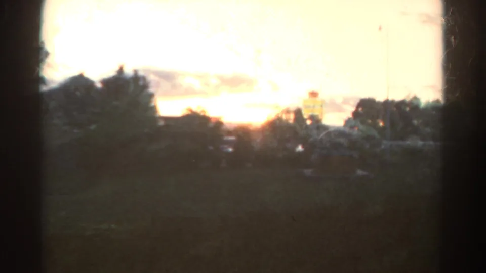 1967 Colorado home-movie frame with warm sunset glare, dark foreground land, tree silhouettes, possible distant roofline, and soft 8mm grain.