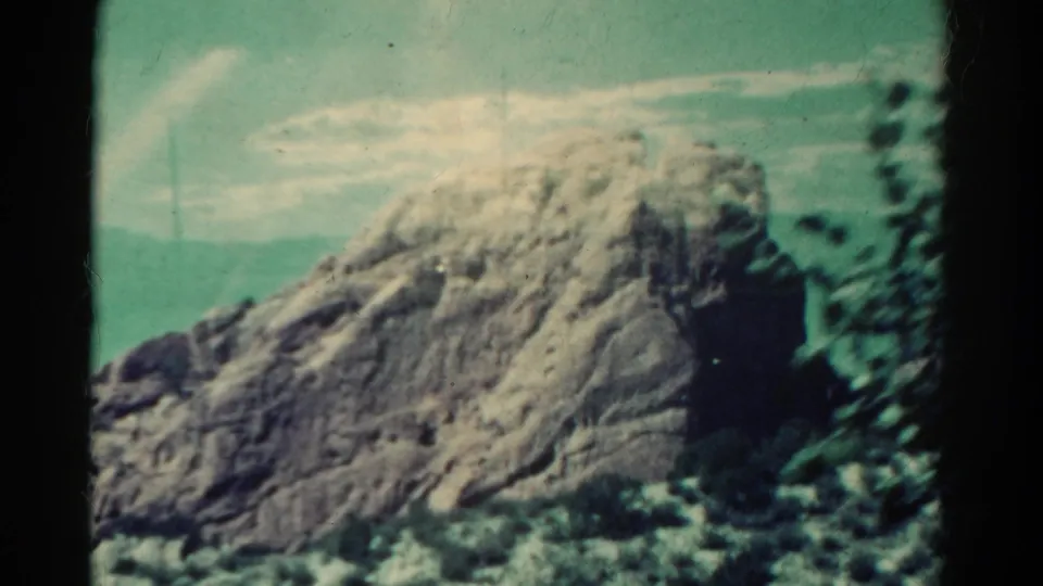 1967 Colorado 8mm frame of a large rock formation with shrubs in the foreground and pale clouds overhead.