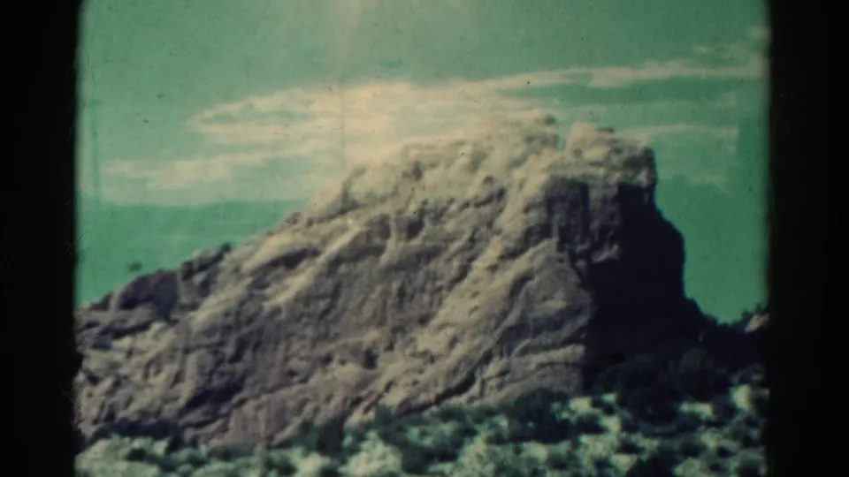1967 Colorado landscape frame showing a rounded rocky cliff, sparse foreground vegetation, and pale clouds in a blue-green sky.