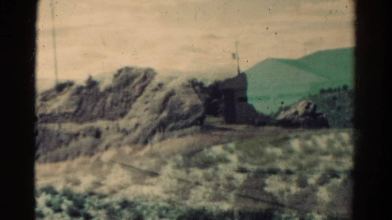 1967 Colorado home-movie frame with rocky terrain, a small hillside structure, flag, utility pole, and distant mountain ridge.