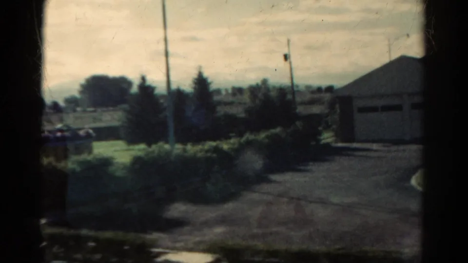 1967 Colorado 8mm frame showing a residential exterior with a driveway, garage-like building, trees, lawn, and utility poles.