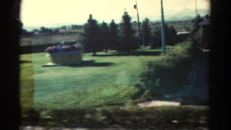 1967 Colorado home-movie frame showing a grassy yard, decorative planter, trees, utility pole, and distant hills.