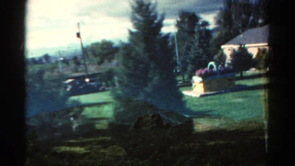 1967 Colorado home-movie view across a suburban lawn toward a parked car, flower planter, trees, and a nearby house.