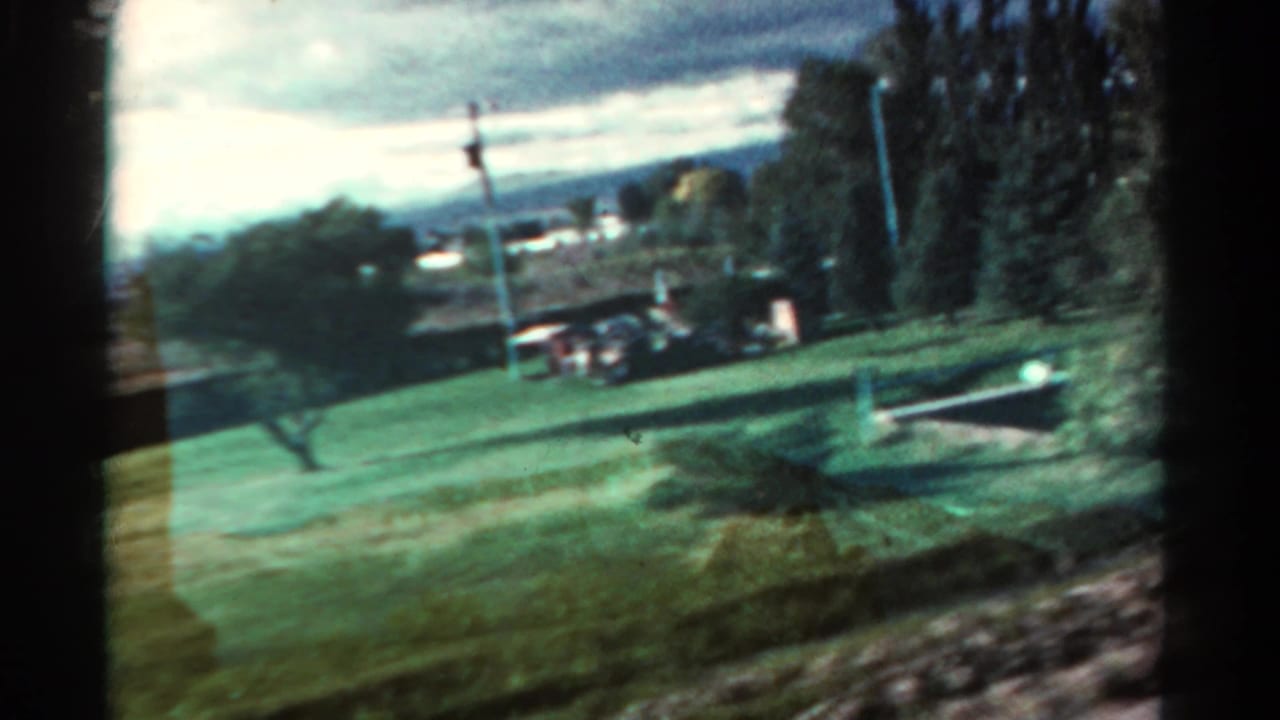 1967 Colorado home-movie frame from a moving vehicle, showing green roadside grounds, trees, poles, small buildings, and distant hills.