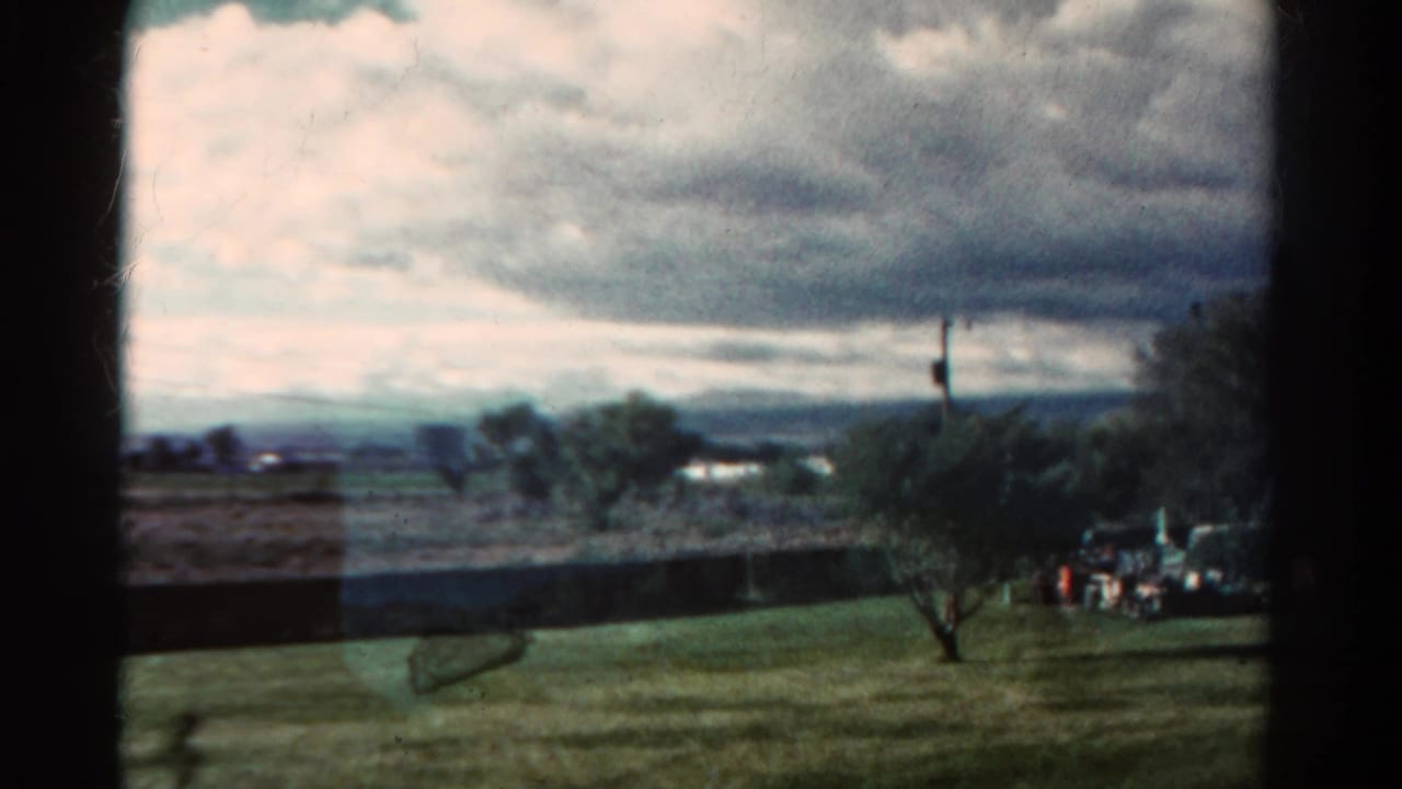 1967 Colorado landscape view with open grass, small trees, distant hills, dramatic clouds, and distant people or objects near the right edge.