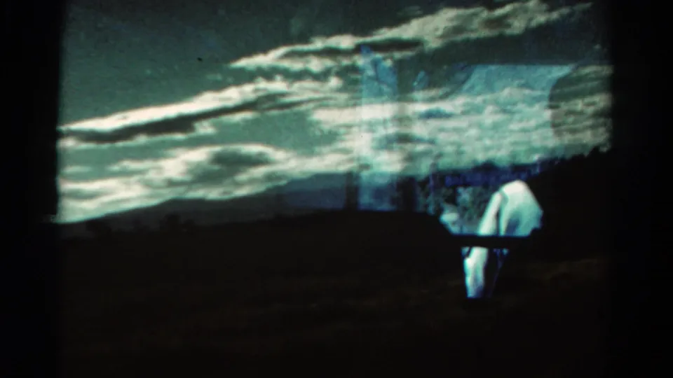 Projected or re-filmed 8mm view showing clouds, dark hill silhouettes, and a pale standing person near the lower right.