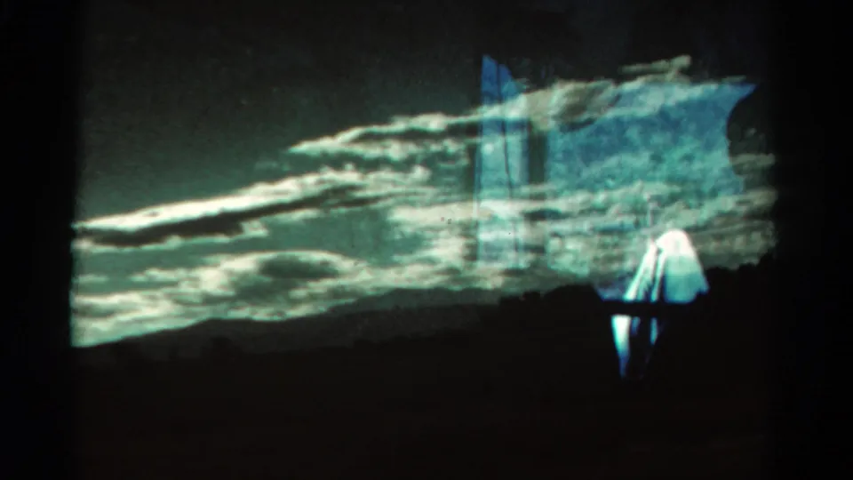 Projected or re-filmed 8mm scene with dramatic cloud bands, dark hills, blue flare, and a partially visible person at lower right.