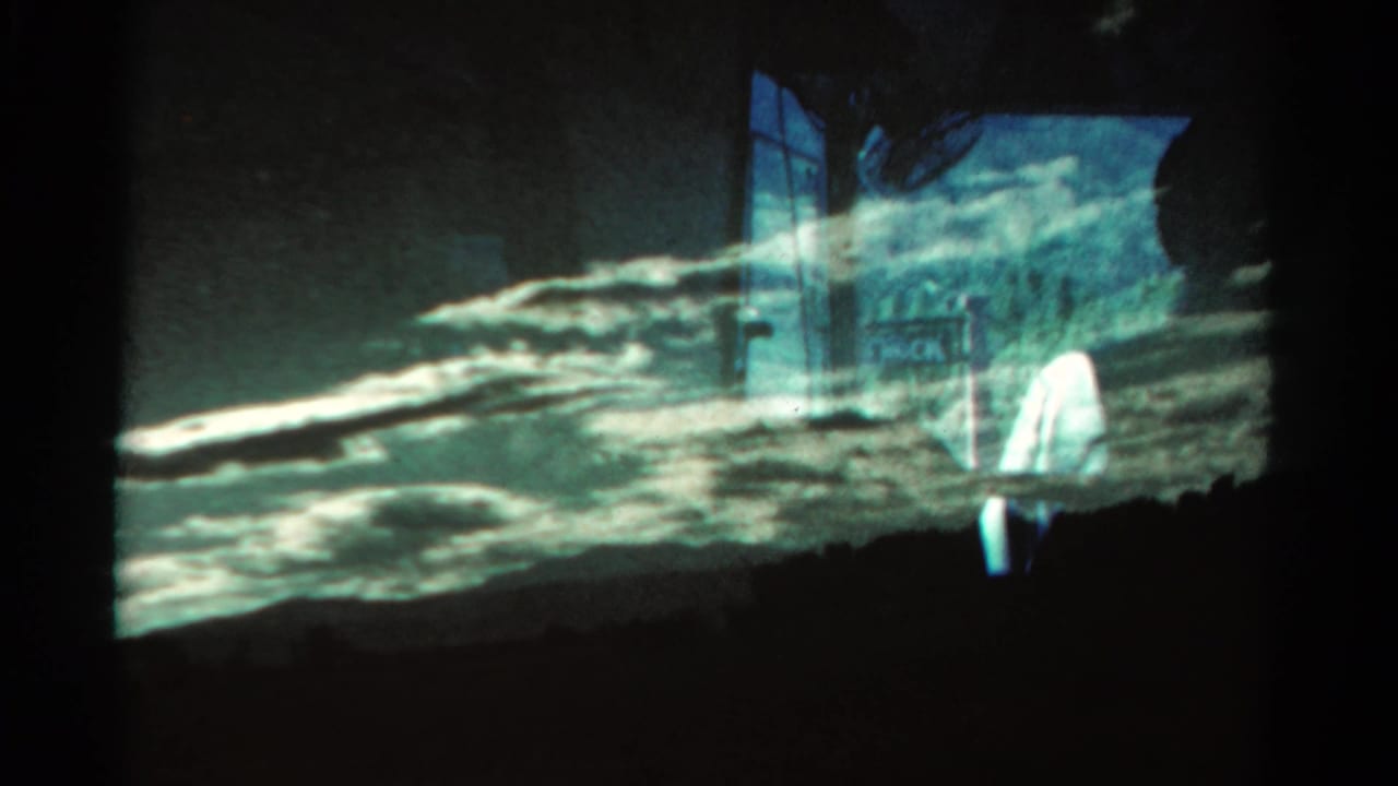 1967 Colorado 8mm frame showing a silhouetted mountain ridge, dramatic cloud band, dark foreground, and a faint pale figure near the roadside.