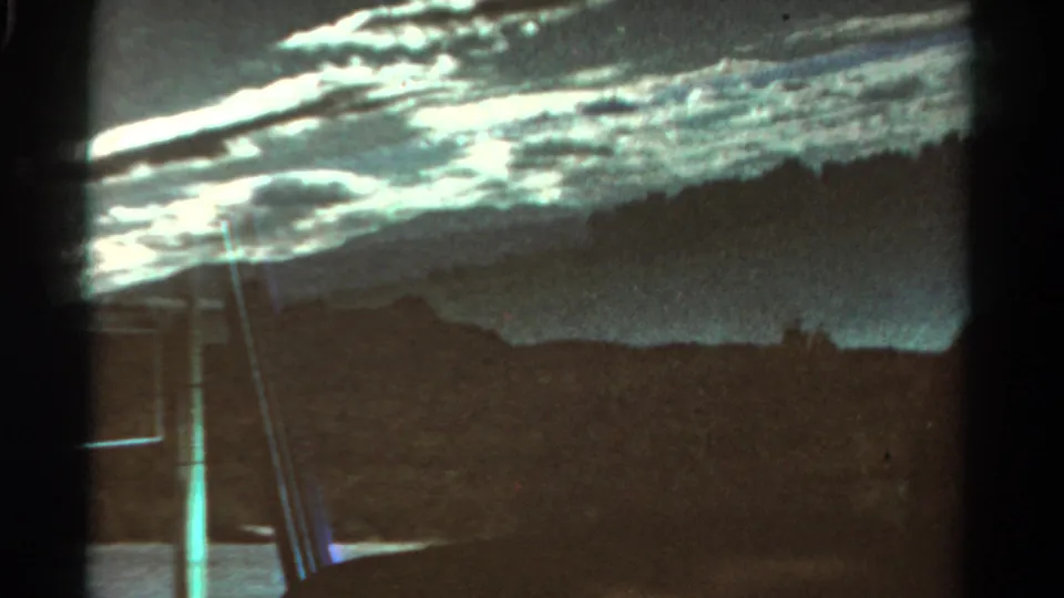 1967 Colorado 8mm frame showing a shadowed mountain horizon, layered clouds, dark passing foreground, and vertical light streaks from filming or reflection.