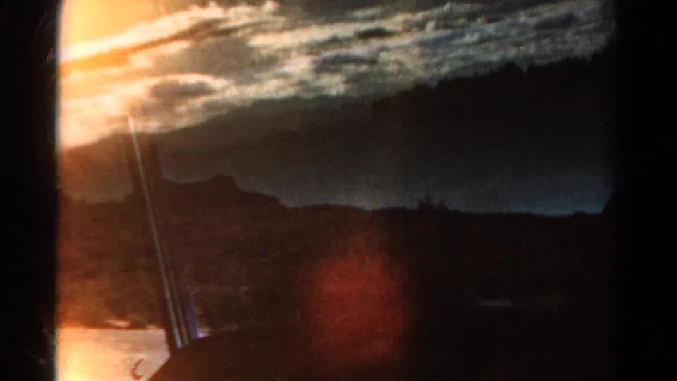 1967 Colorado 8mm frame with silhouetted mountain scenery, streaked clouds, dark foreground, and a warm orange-red light leak or flare.
