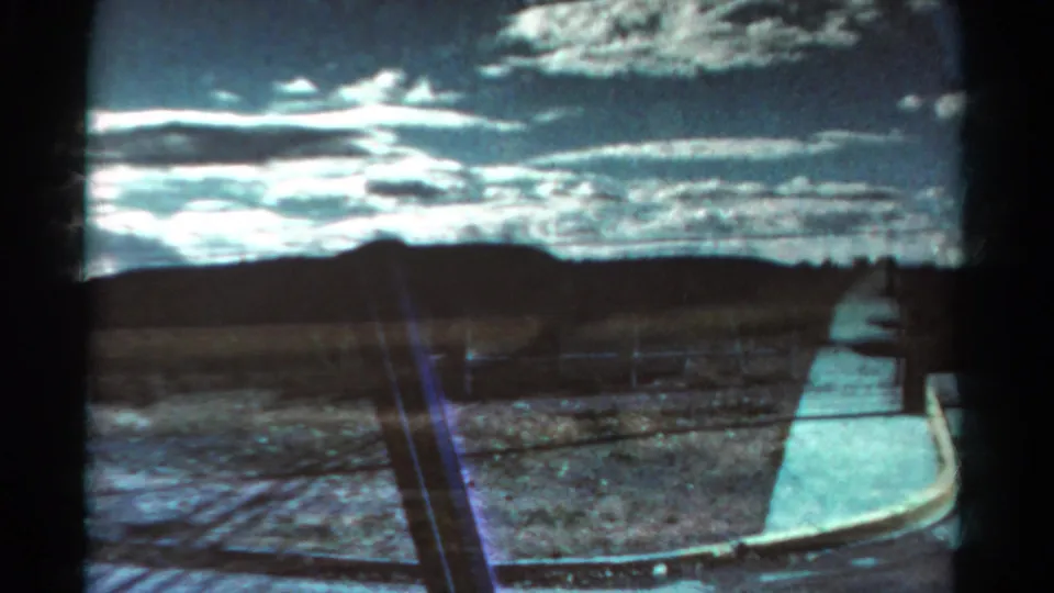 Final 1967 Colorado frame shows roadside shoulder and open rural ground under a bright clouded sky, with window framing and film artifacts visible.